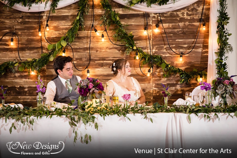 A bride and groom sit in front of a rustic barnwood backdrop striped with wild garland, at a head table decorated with garland, and bud vases of various heights sporting Deep and light purple flowers.